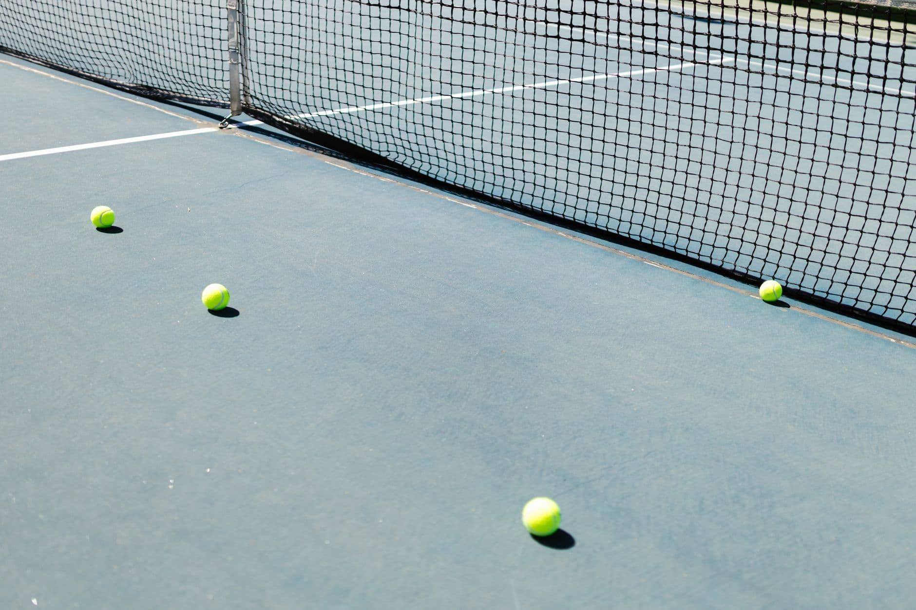 Un court de tennis avec un filet et plusieurs balles de tennis au sol, bien éclairé par la lumière du soleil.